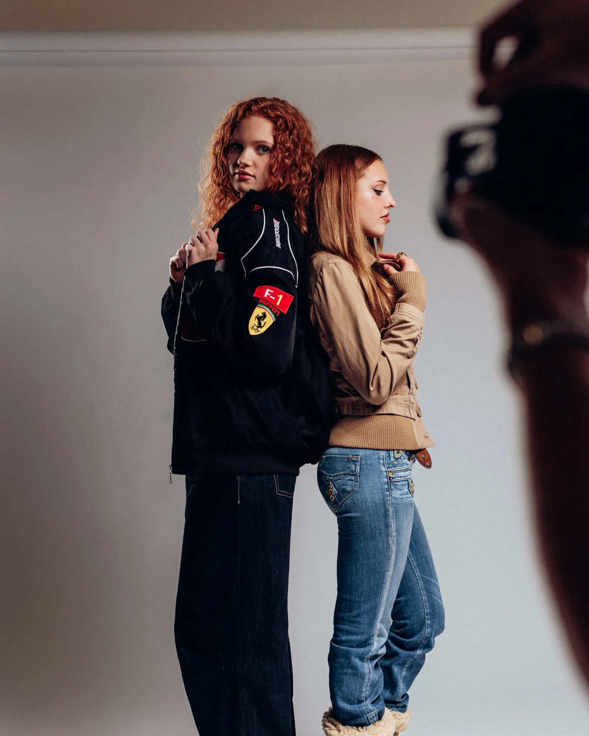 Two young women posing back-to-back in a studio; one in a black Ferrari F1 jacket, the other in a tan jacket.