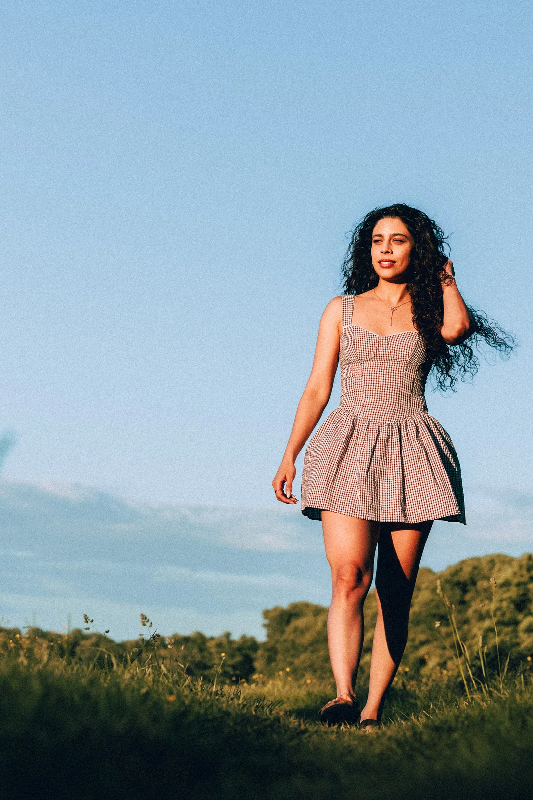 Woman with curly hair in a red gingham dress standing in a sunny grassy field.