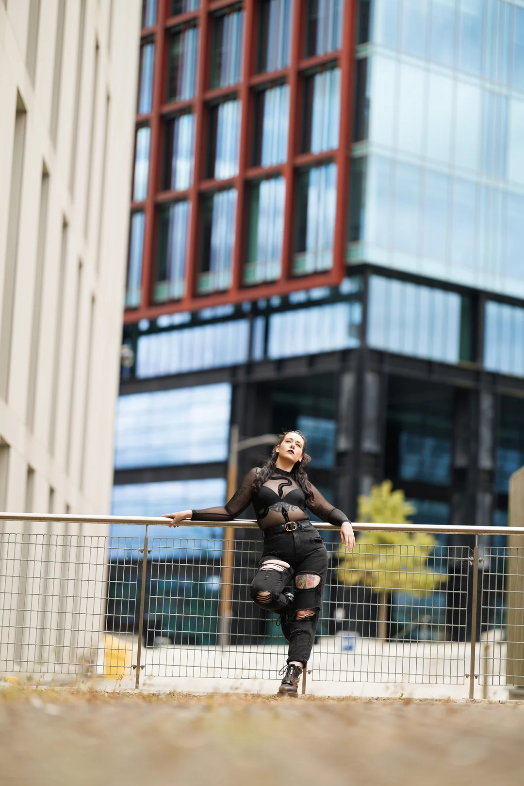Woman in a sheer black top and ripped jeans posing by a railing in an urban setting.