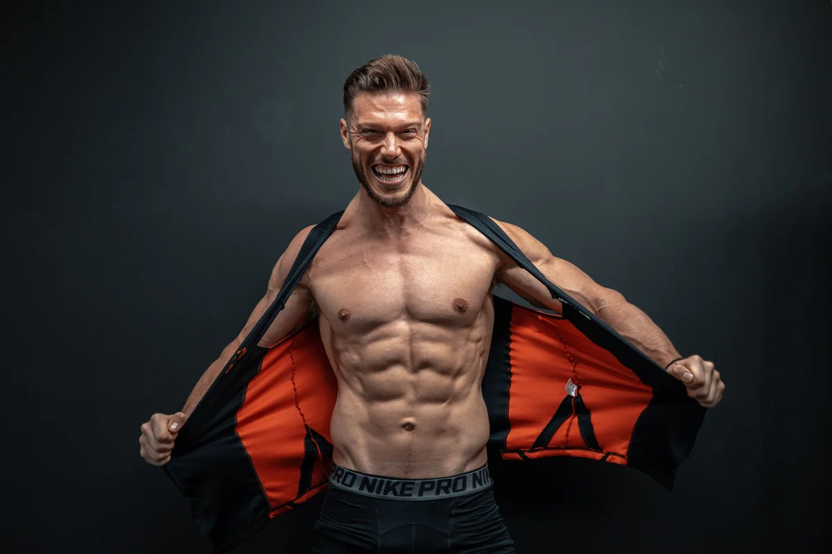Muscular man with a beard smiling and opening a black and orange jacket to show his abs against a dark background.