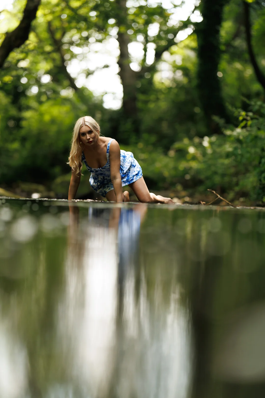 A blonde woman in a blue patterned dress posing by a reflective body of water in a lush green forest.