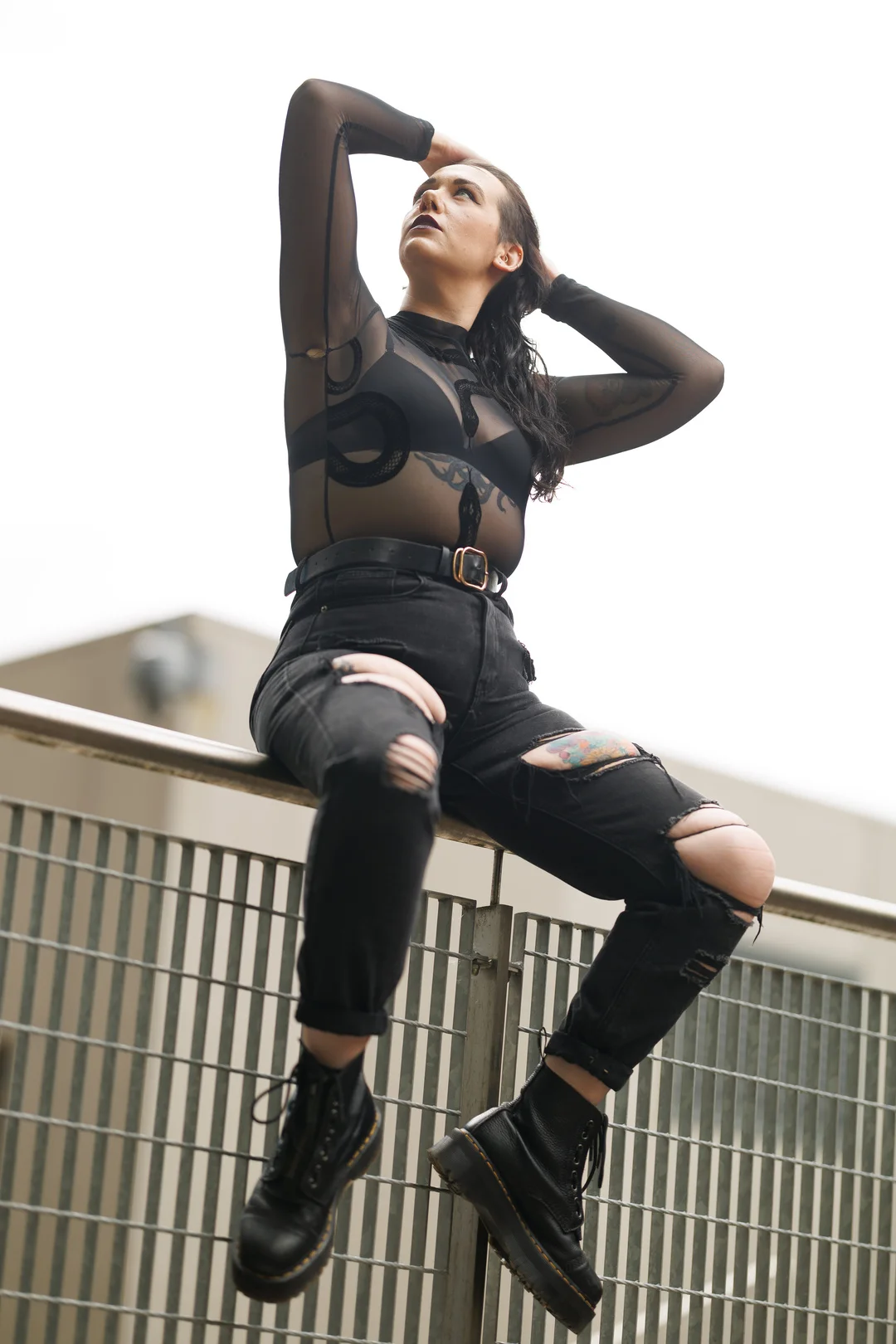 Full-body shot of a person in a sheer black top and ripped black jeans posing on an urban metal railing.