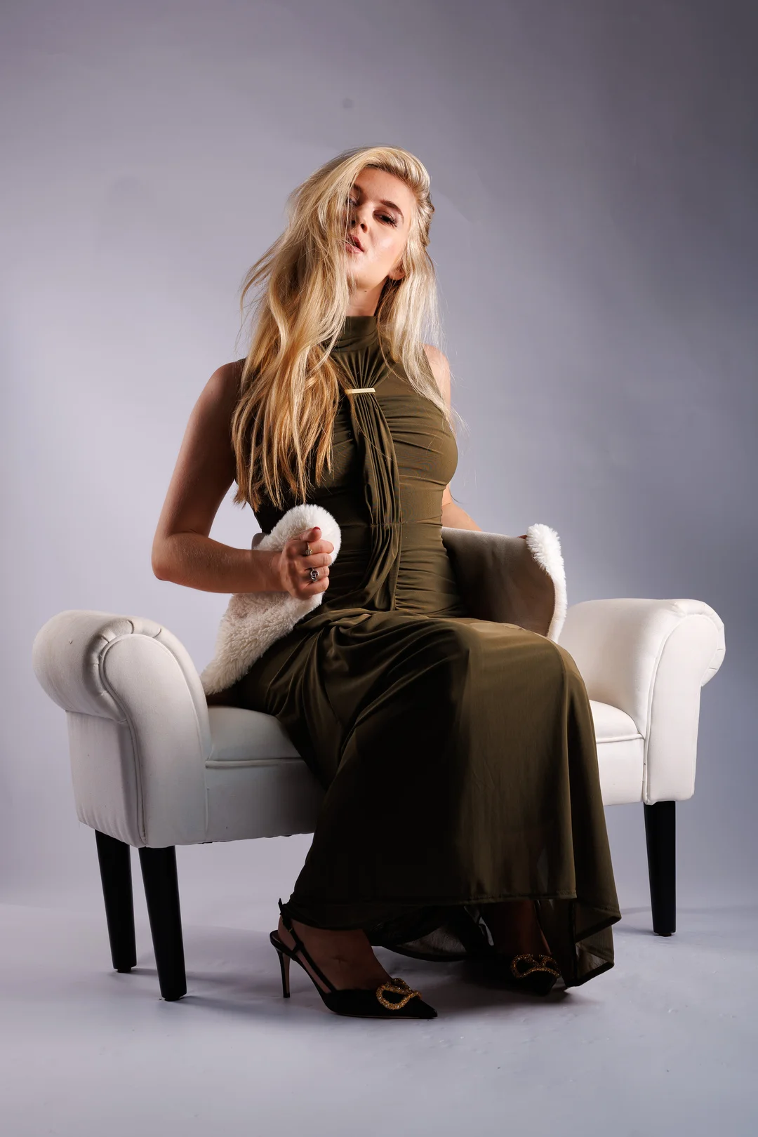 Blonde woman in an olive green gown posing on a white armchair in a studio setting.