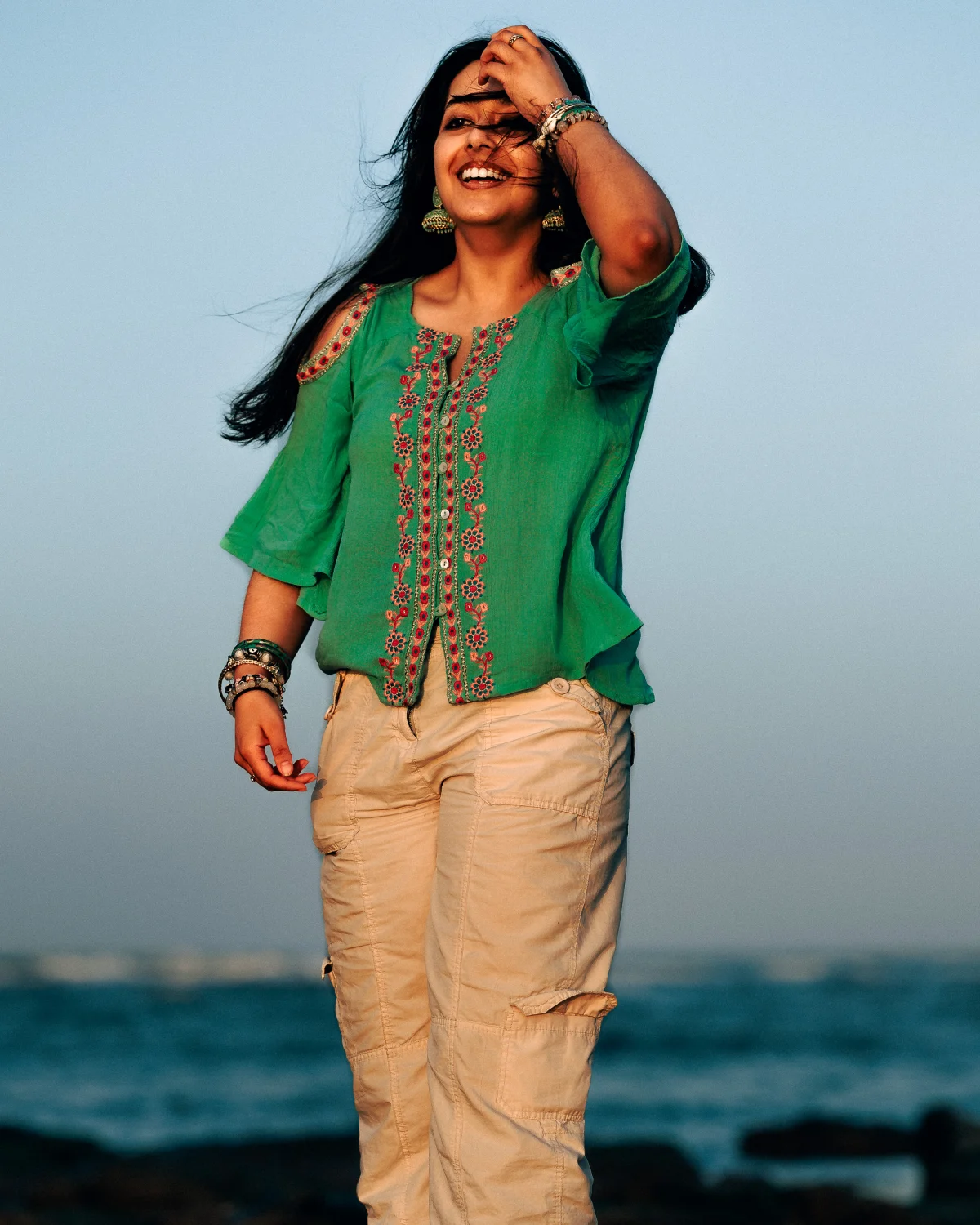 Smiling woman in a green embroidered top and khaki pants on a beach at dusk.