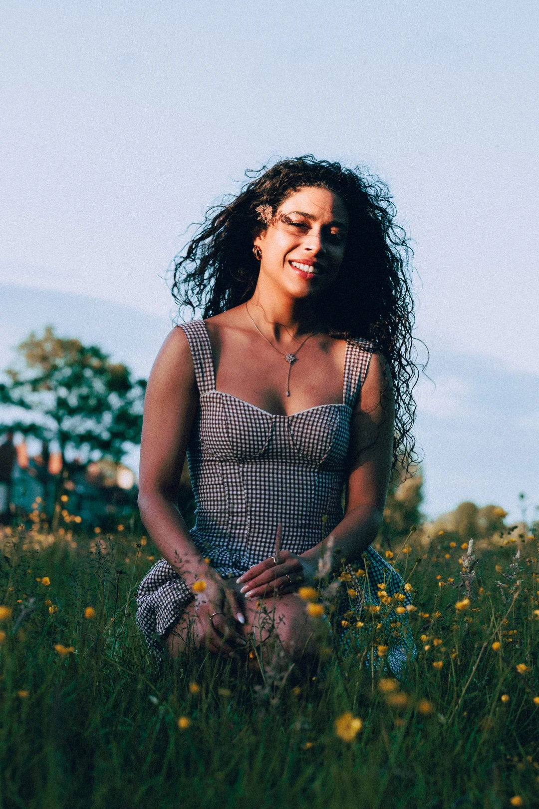 A smiling woman with curly hair in a gingham dress, kneeling in a field of yellow wildflowers at sunset.