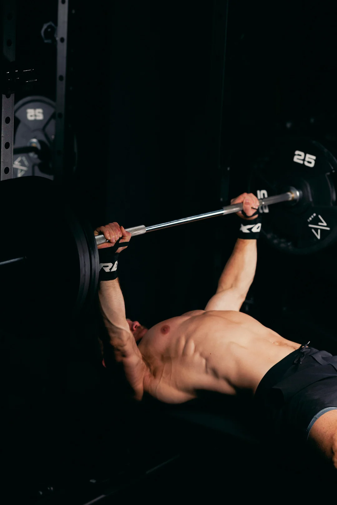 A shirtless man performing a bench press in a dark gym, highlighting muscle definition.