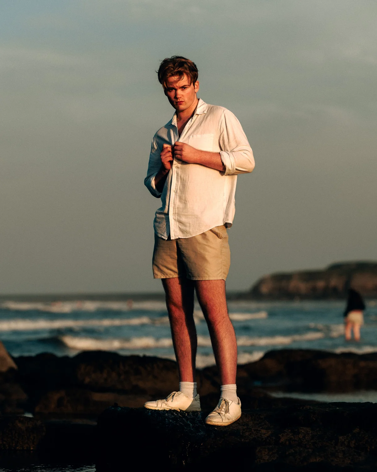 Full-body portrait of a young man in a white linen shirt and khaki shorts standing on a rocky beach at dusk.