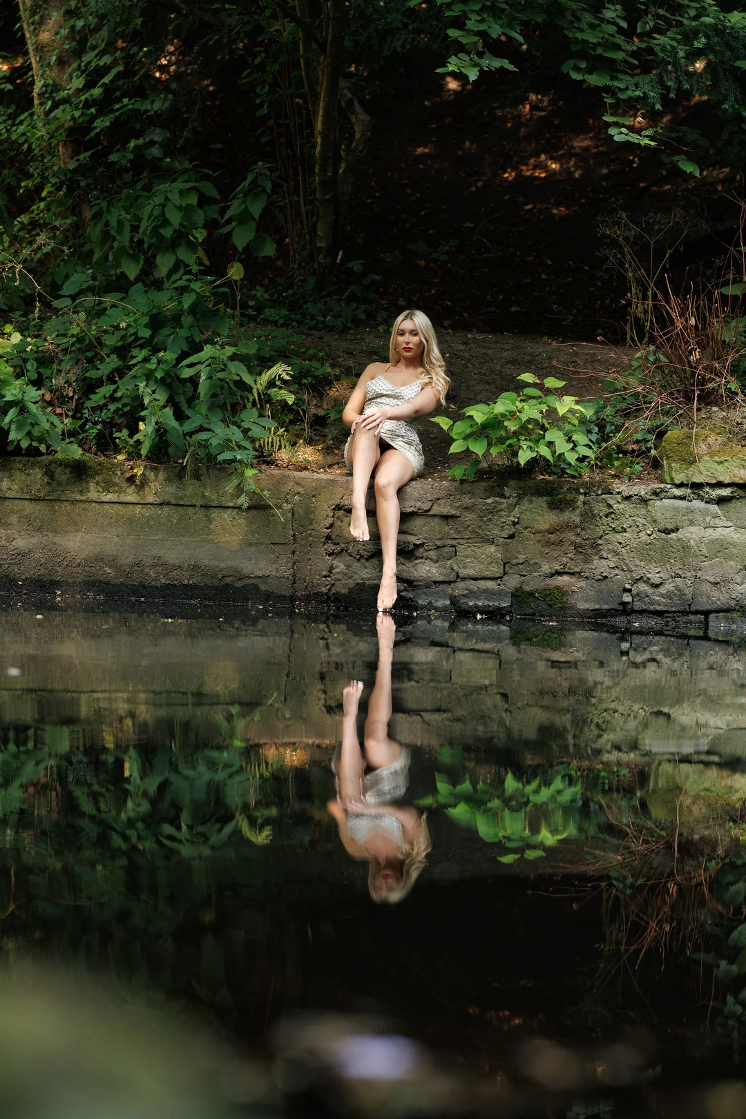 Blonde woman in a short silver dress sitting on a stone wall by a pond, reflecting in the water.