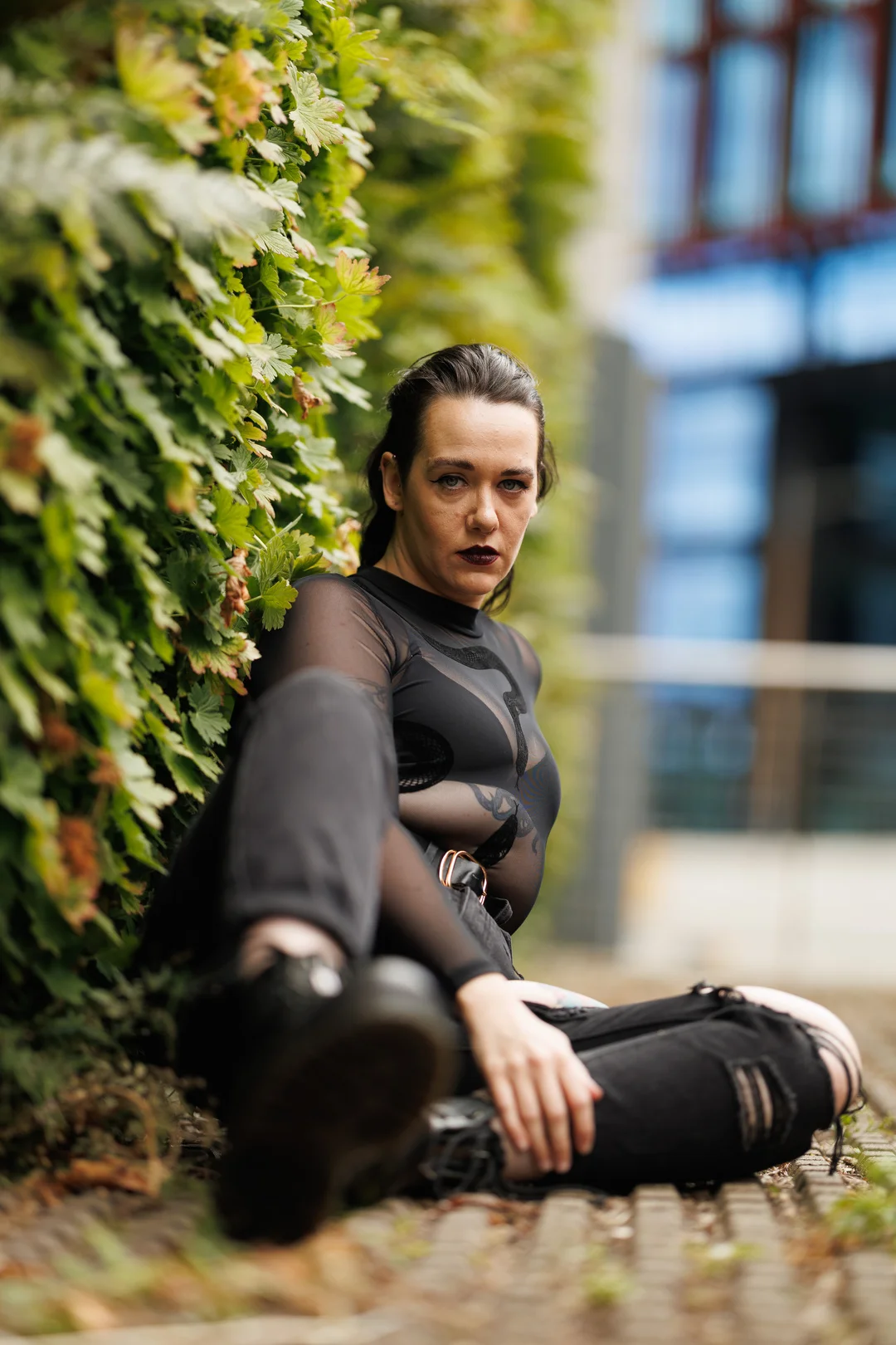 A woman with dark makeup and a sheer black top sitting against a lush green plant wall outdoors.