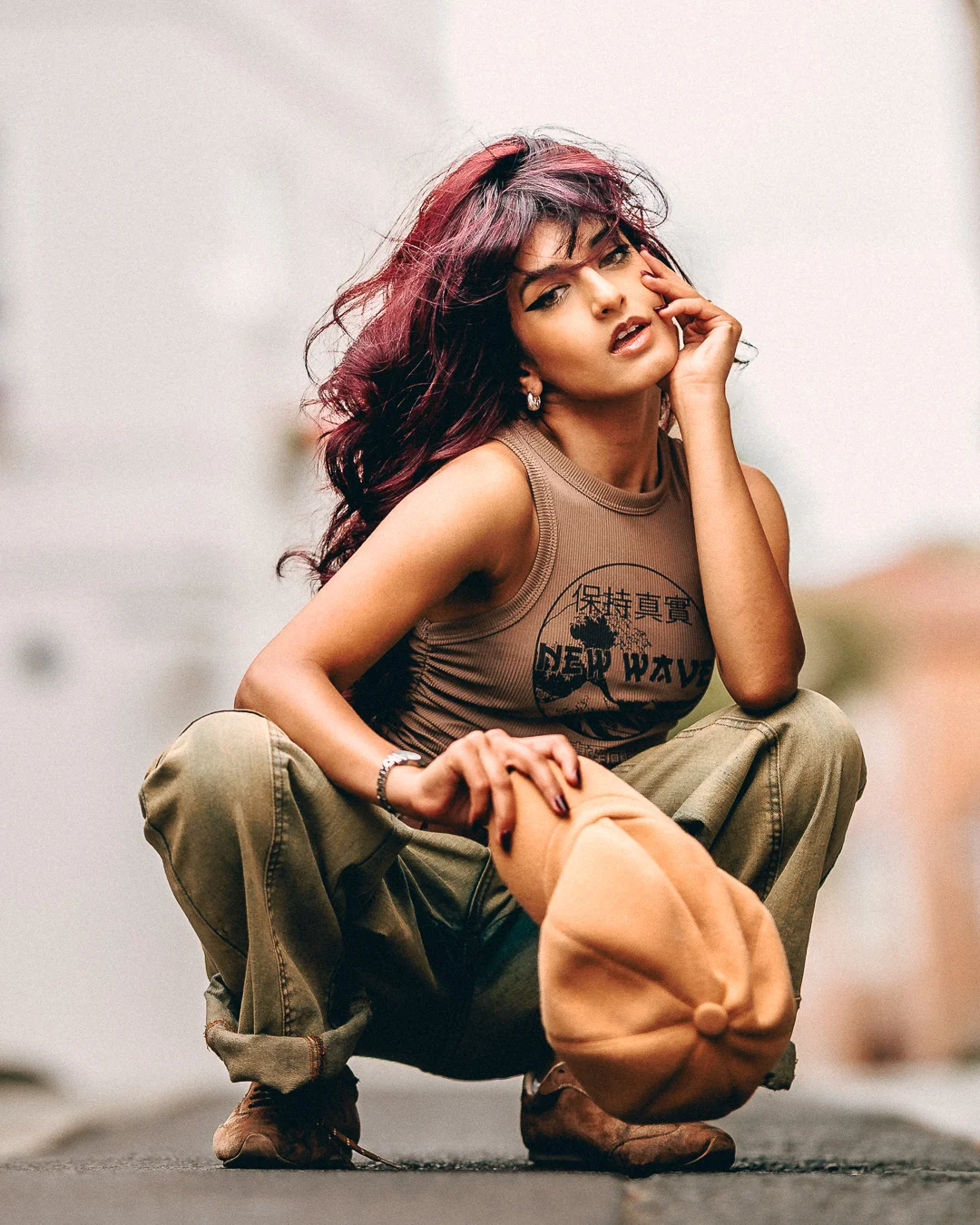 A woman with reddish-purple hair crouching in an urban setting, wearing a tan tank top and cargo pants.