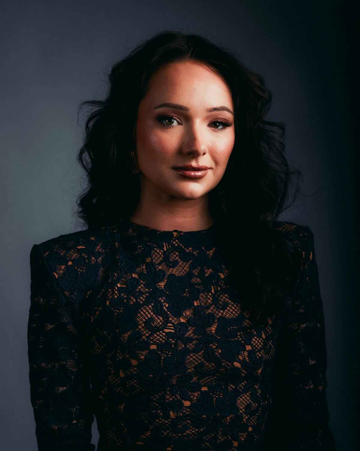 Woman with long dark wavy hair in a black lace dress posing against a dark grey studio background.