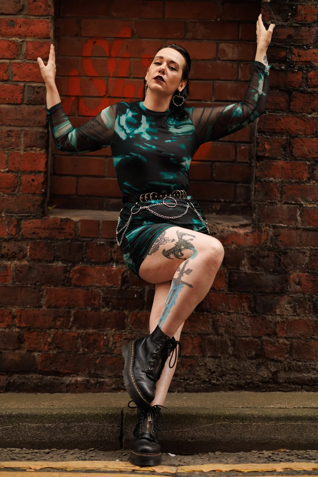 Person in a teal and black dress and black boots posing against a red brick wall.