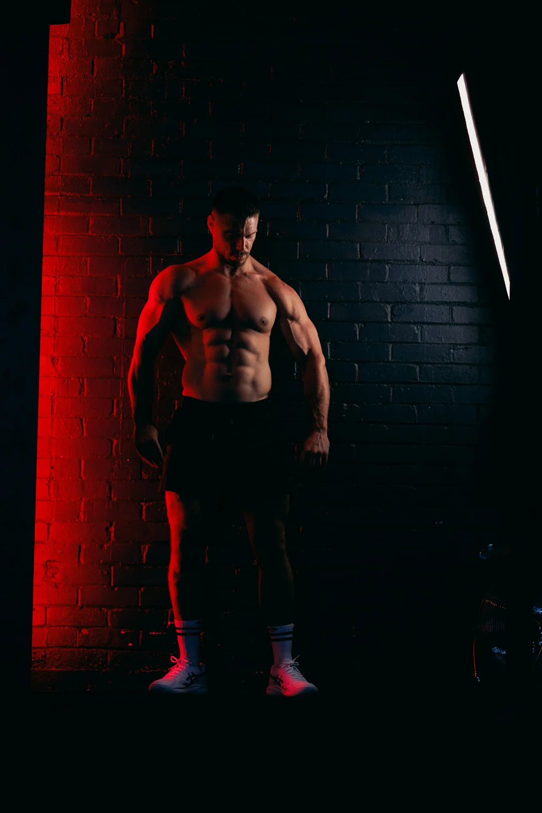 Muscular man posing shirtless against a dark brick wall with dramatic red and white lighting.