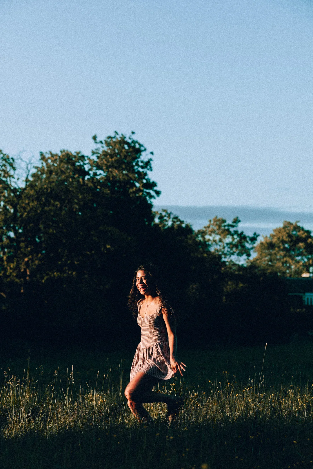 A smiling woman in a red and white checkered dress dancing in a grassy field during golden hour.