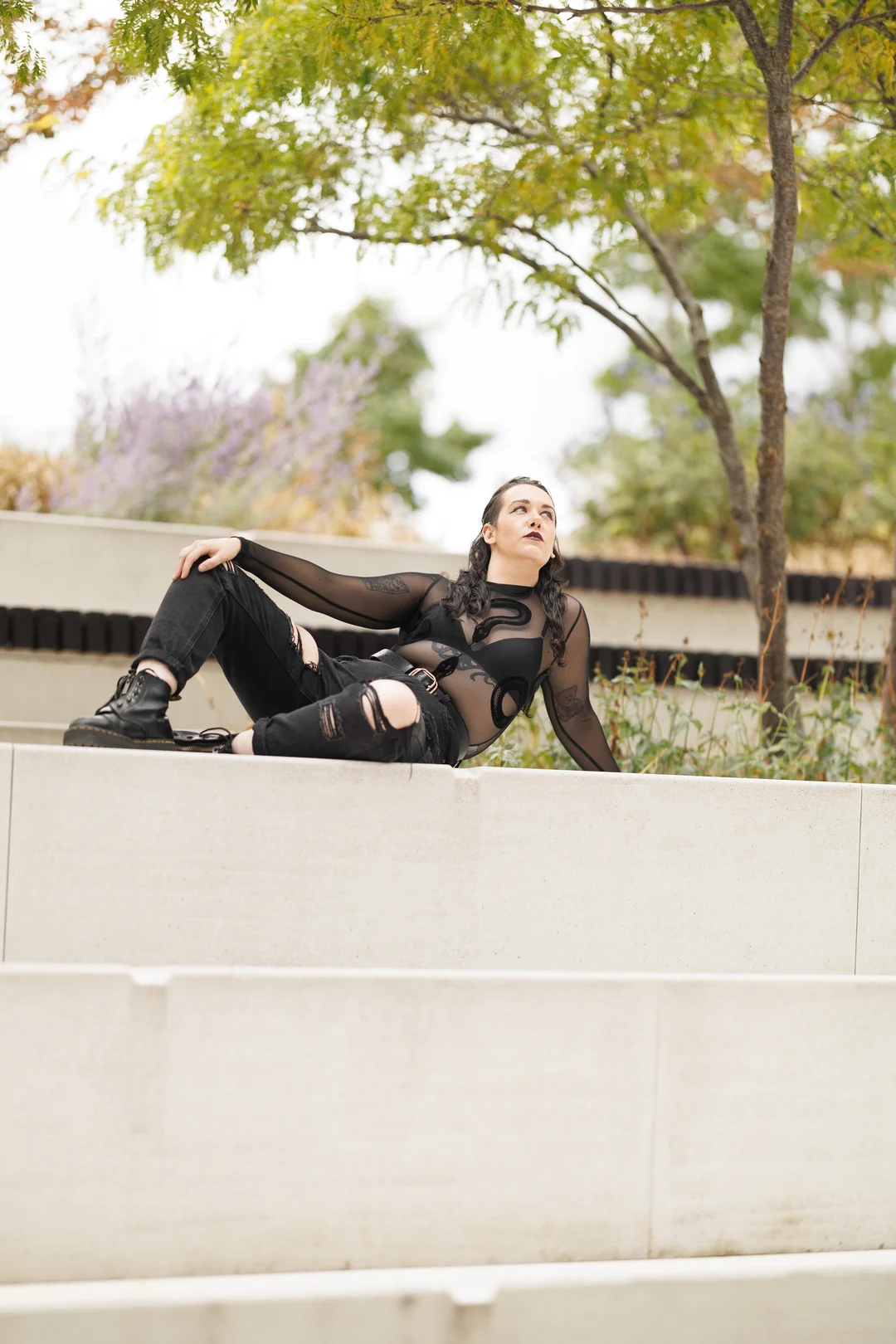 Person in a black sheer top and ripped jeans reclining on concrete steps in an outdoor park setting.