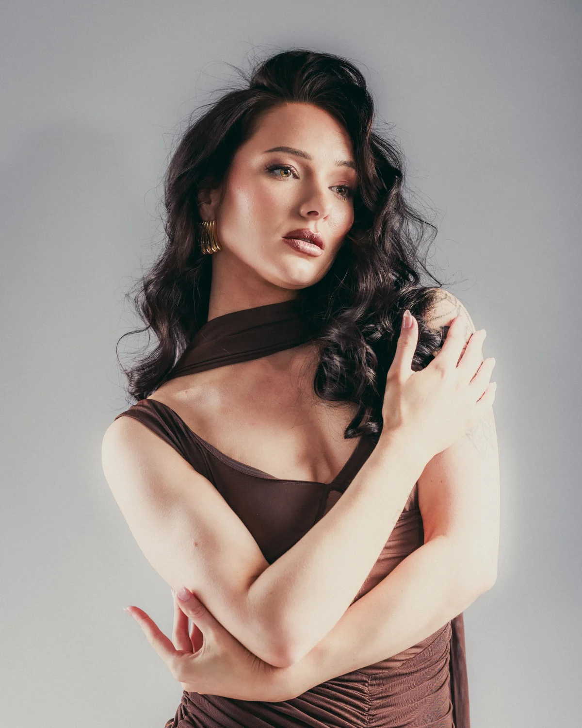 Woman with long dark wavy hair in a brown dress posing against a grey studio background.