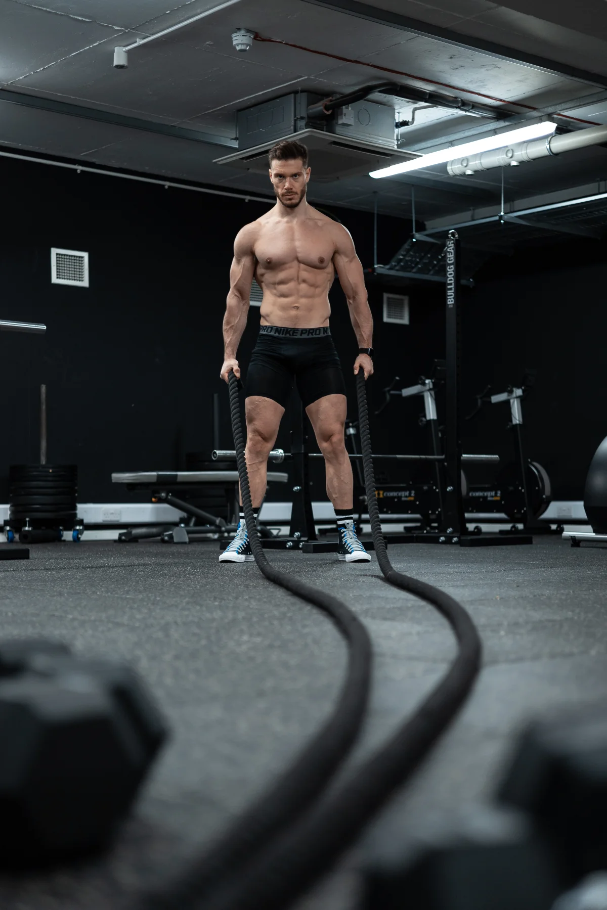 Muscular man in black Nike Pro shorts holding battle ropes in a moody gym setting.