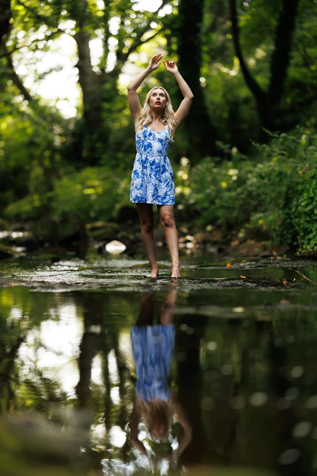 A woman in a blue floral dress posing in a forest stream with her reflection in the water.