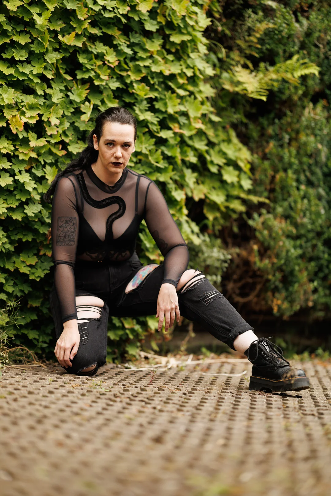 A person with dark hair and makeup in a black mesh top and ripped jeans posing against a green hedge.
