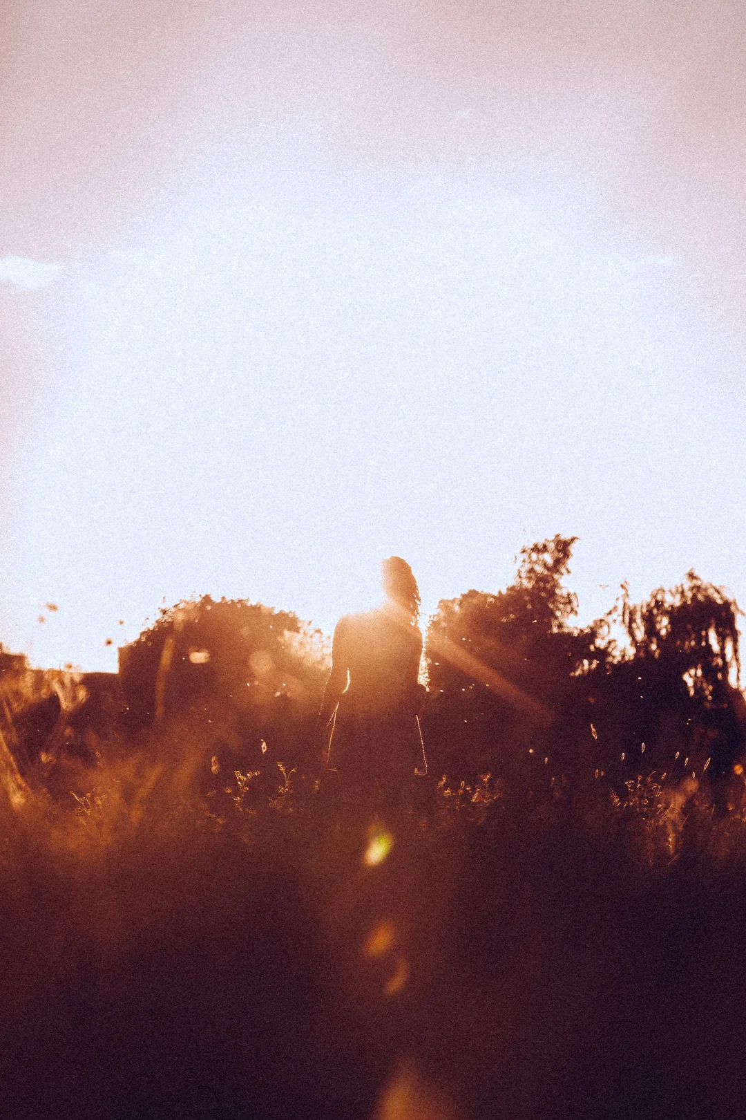 Silhouette of a person standing in a grassy field against a bright, hazy sunset with lens flare.