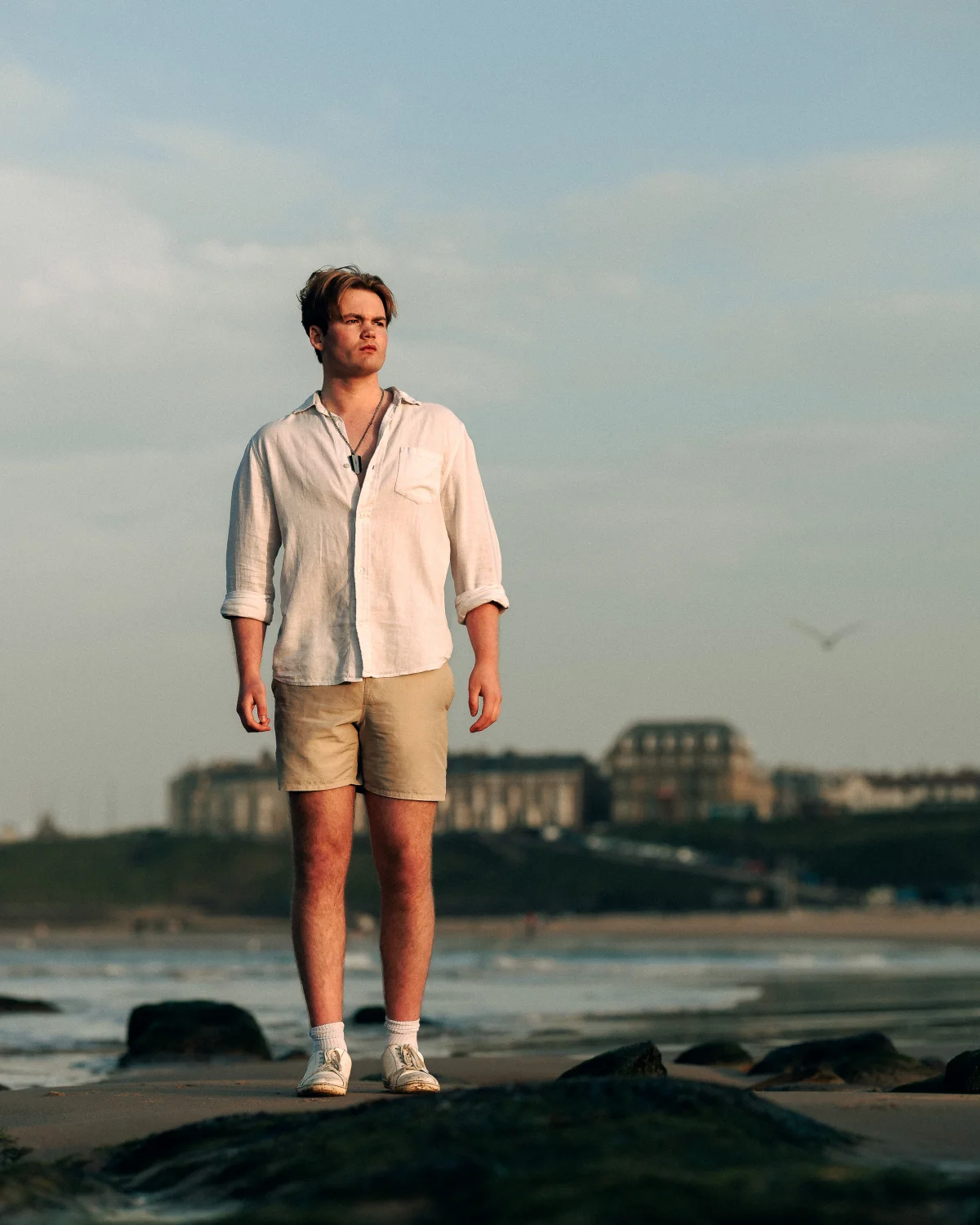 Full-body portrait of a young man in a white linen shirt and beige shorts standing on a beach at dusk.