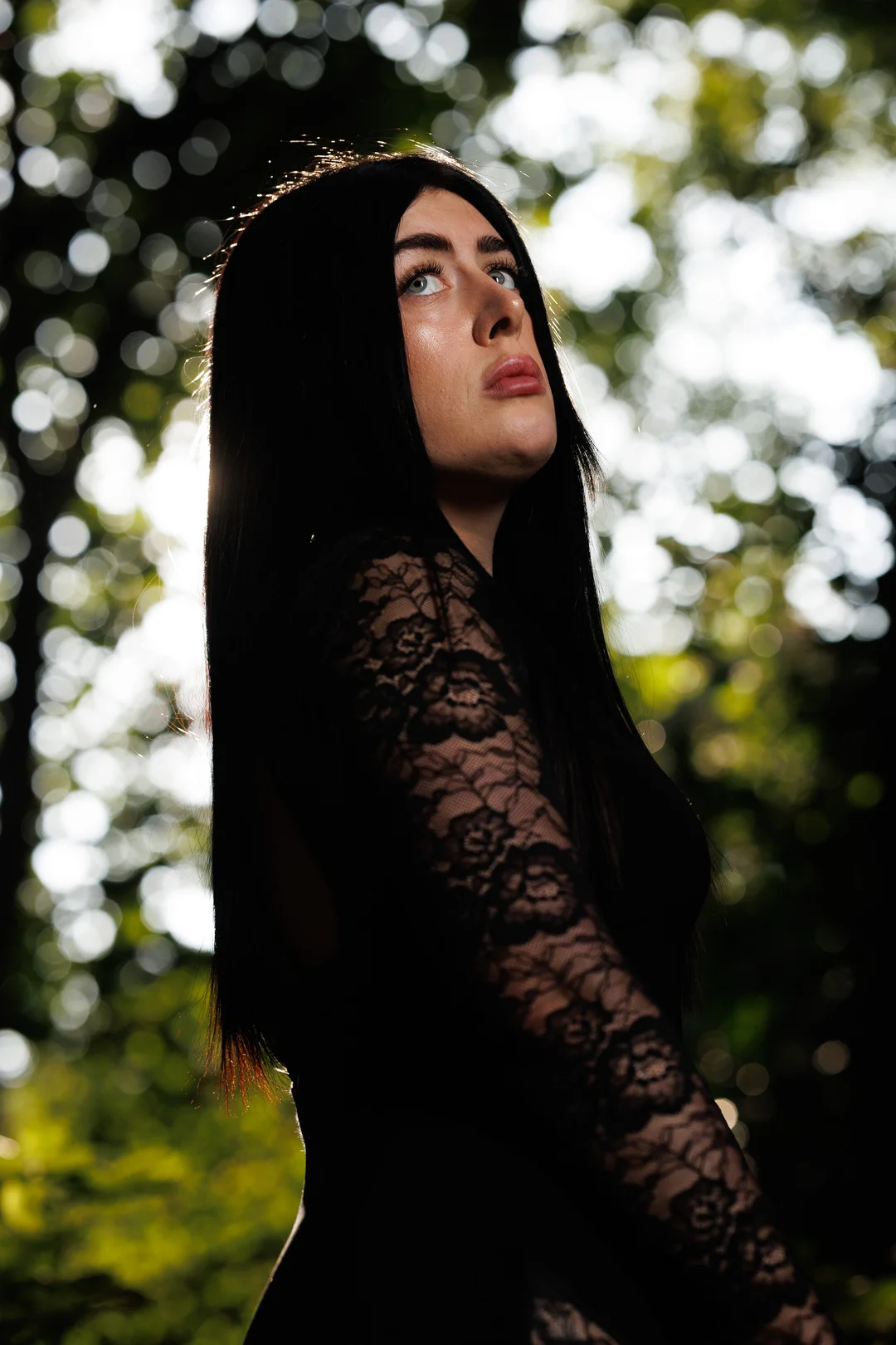 Woman with long black hair in a black lace dress looking up, set against a blurred forest background.
