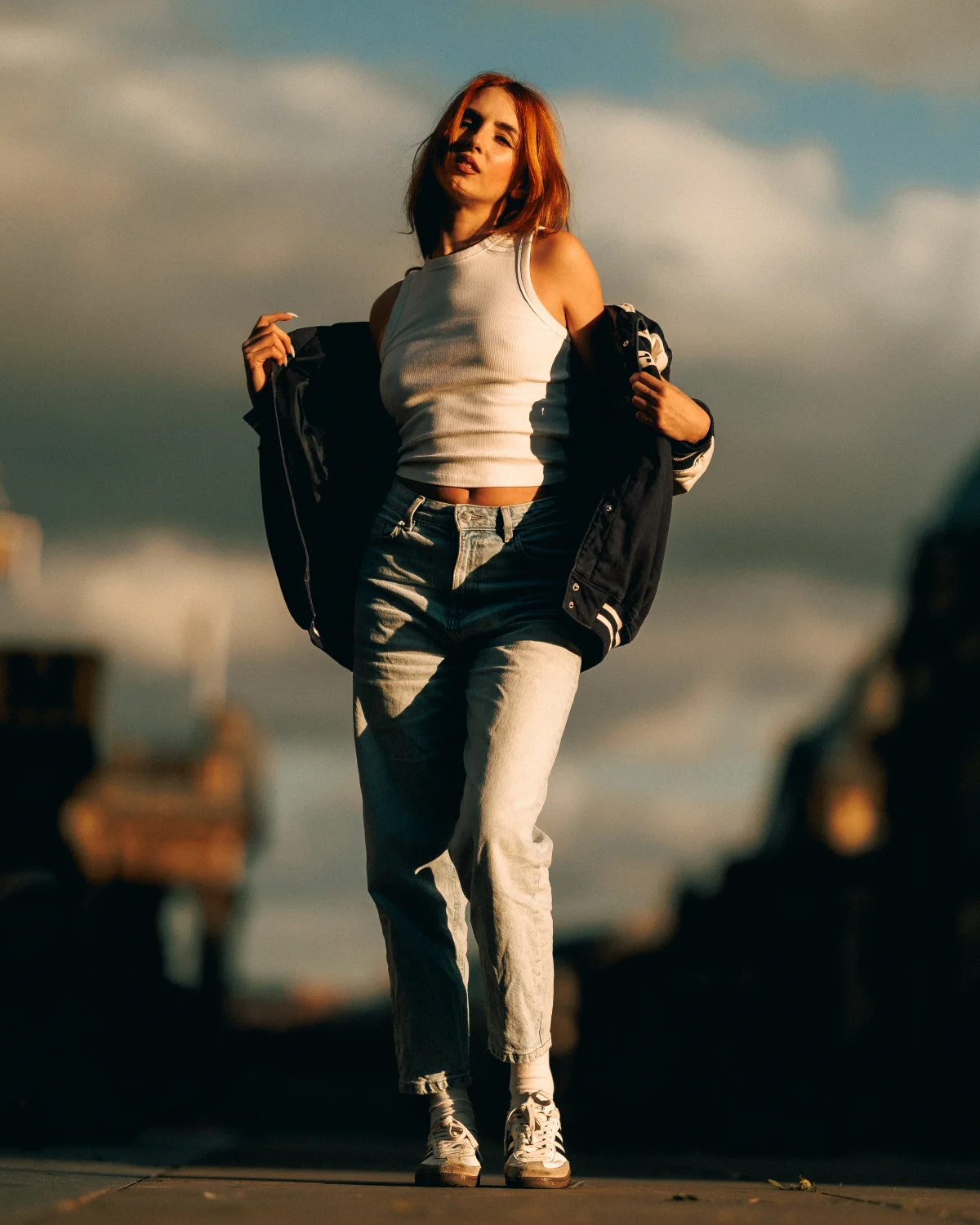 Woman with red hair posing in a white tank top and jeans with a jacket, set against a blurred outdoor background.