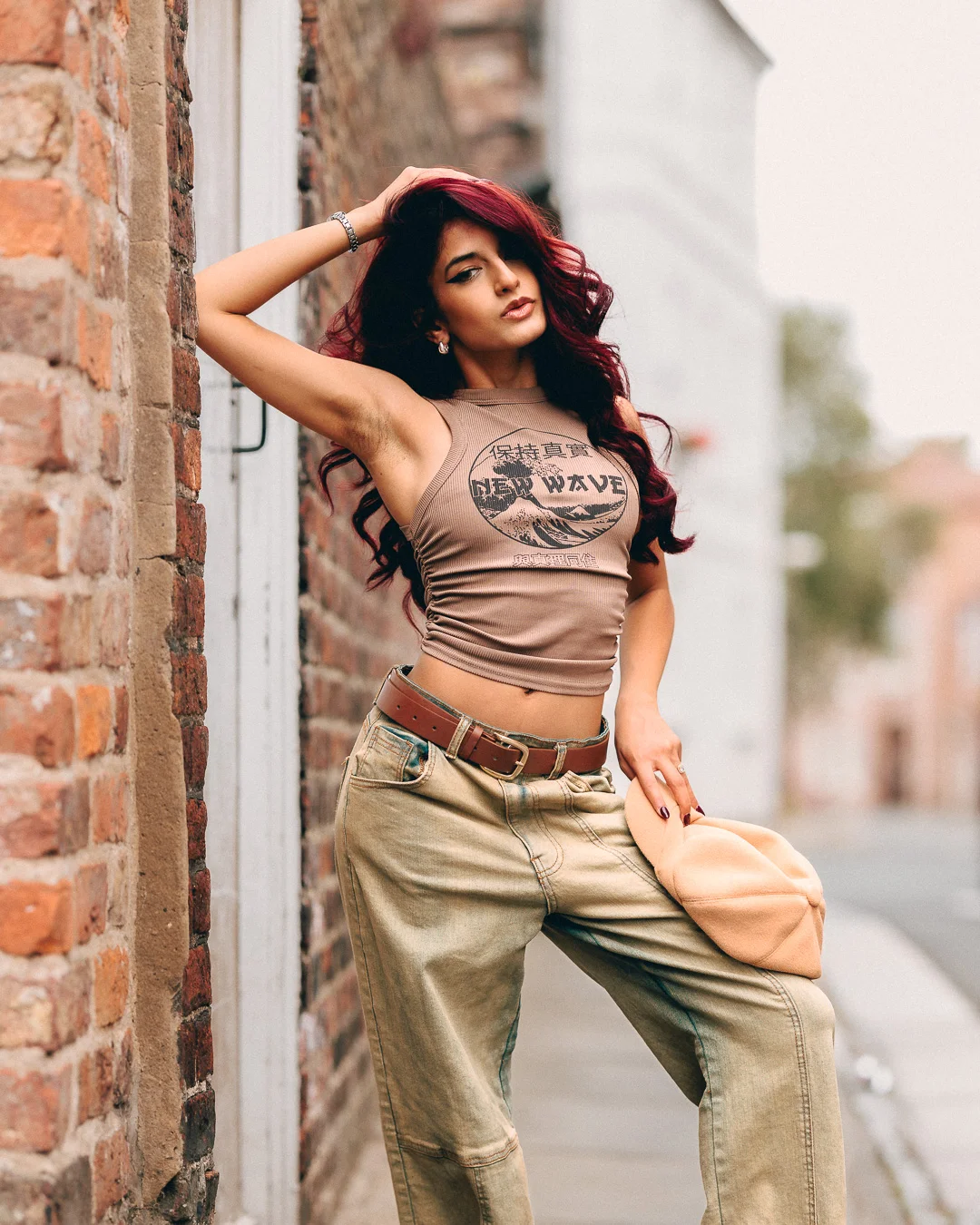 Woman with red hair posing against a brick wall in an urban setting, wearing a tan crop top and khaki pants.