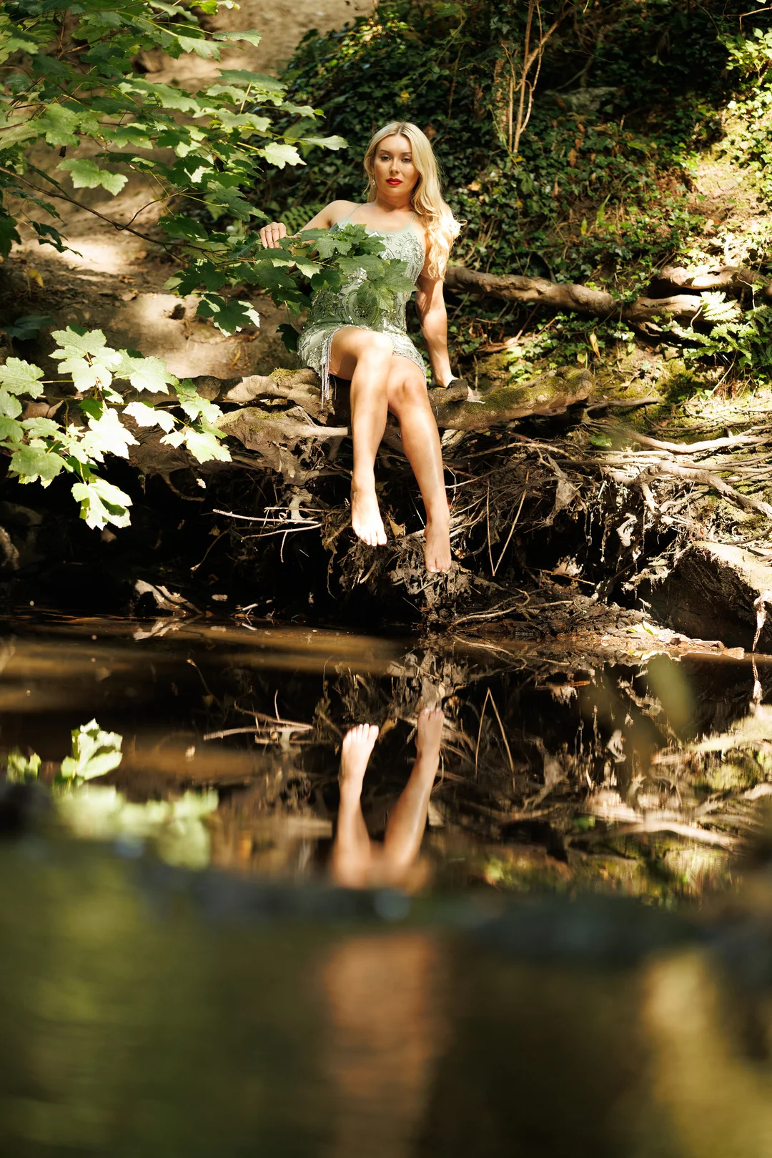 Blonde woman in a sparkly green dress sitting on a log by a stream with a clear reflection in the water.