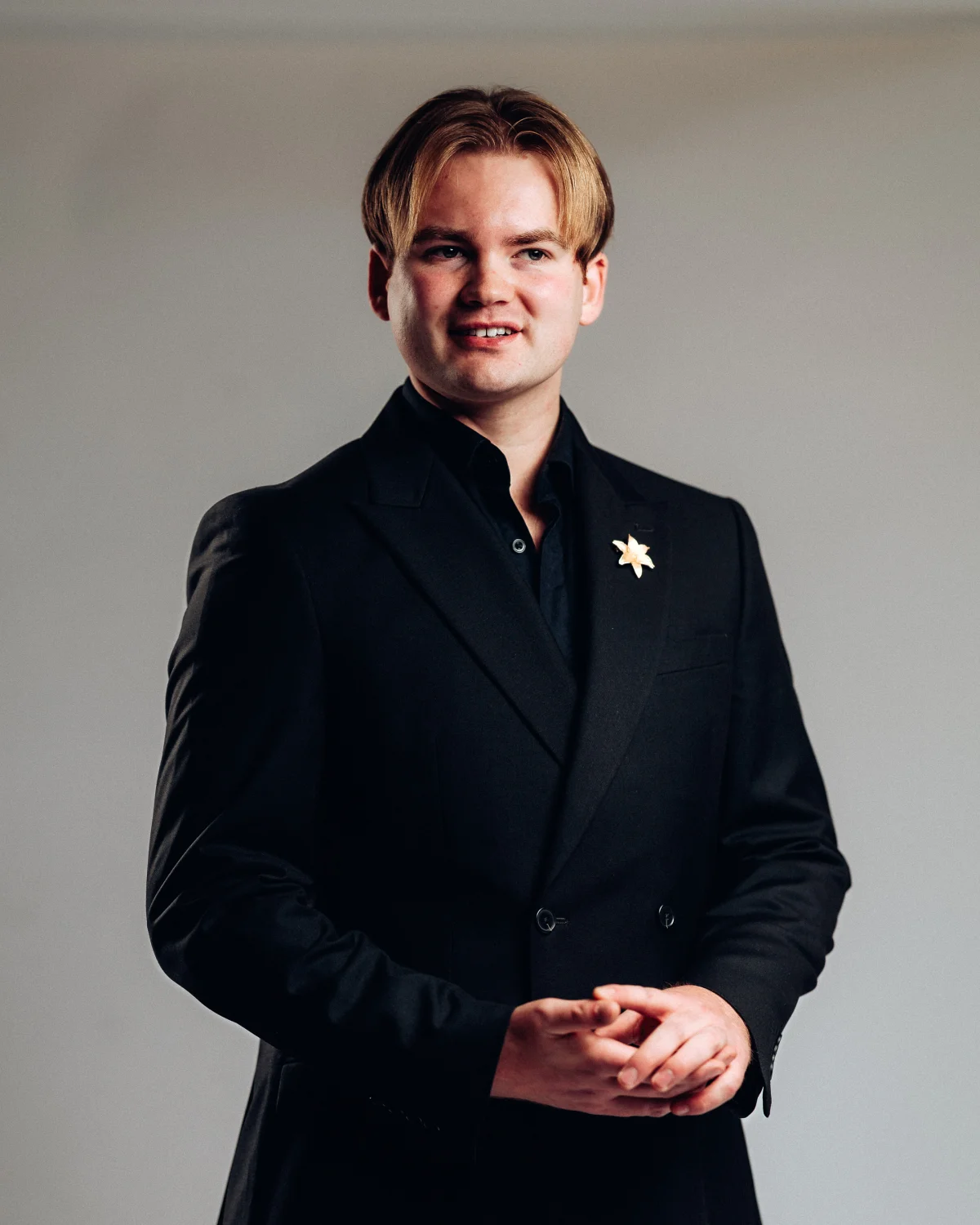 A young man with blond hair in a black suit and shirt posing against a neutral grey studio background.