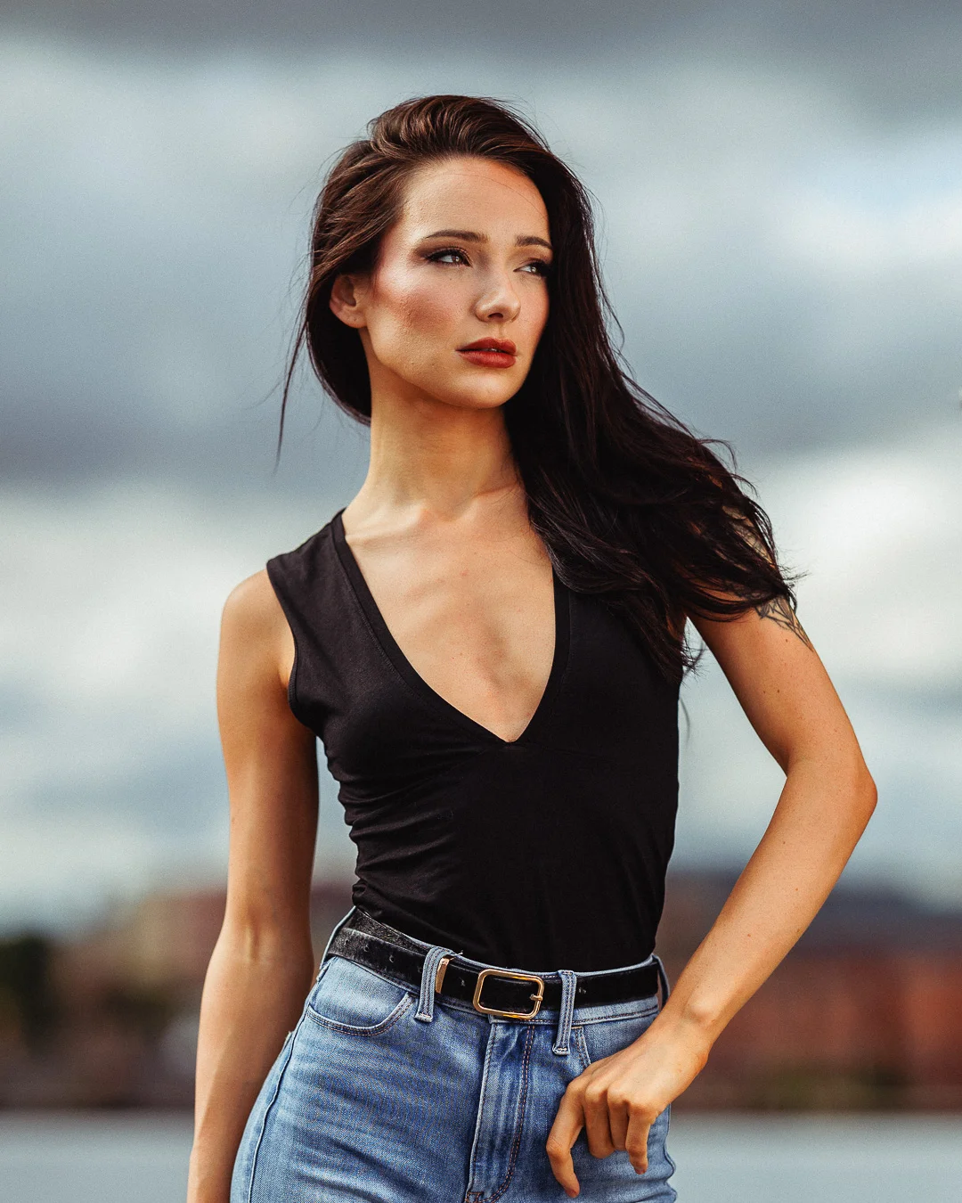 Portrait of a woman with long dark hair in a black V-neck top and blue jeans against a cloudy sky.