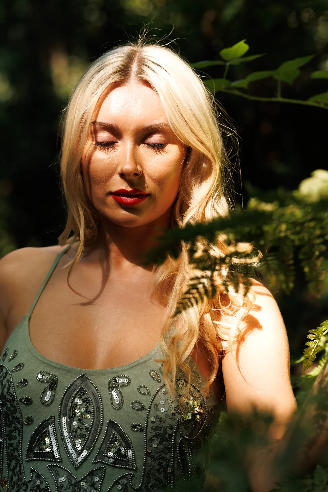 Close-up portrait of a blonde woman with red lipstick and closed eyes in a sunlit forest.