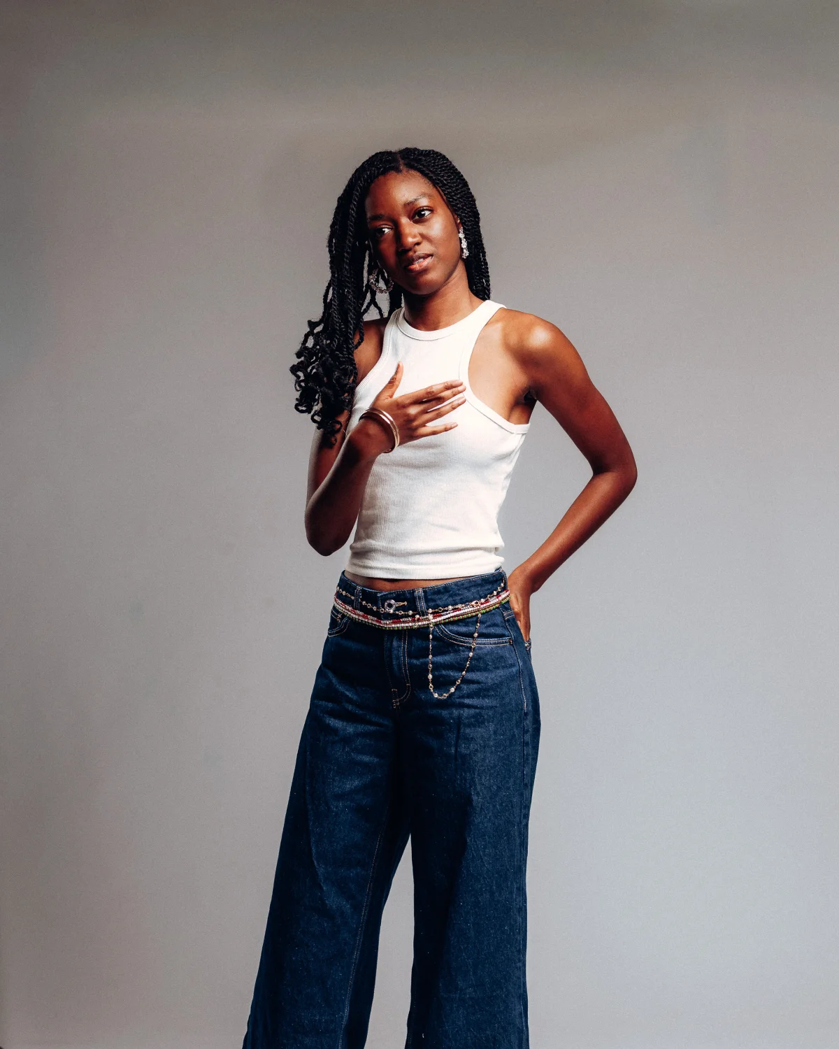 A young Black woman with long braids posing in a white tank top and wide-leg blue jeans against a grey studio background.