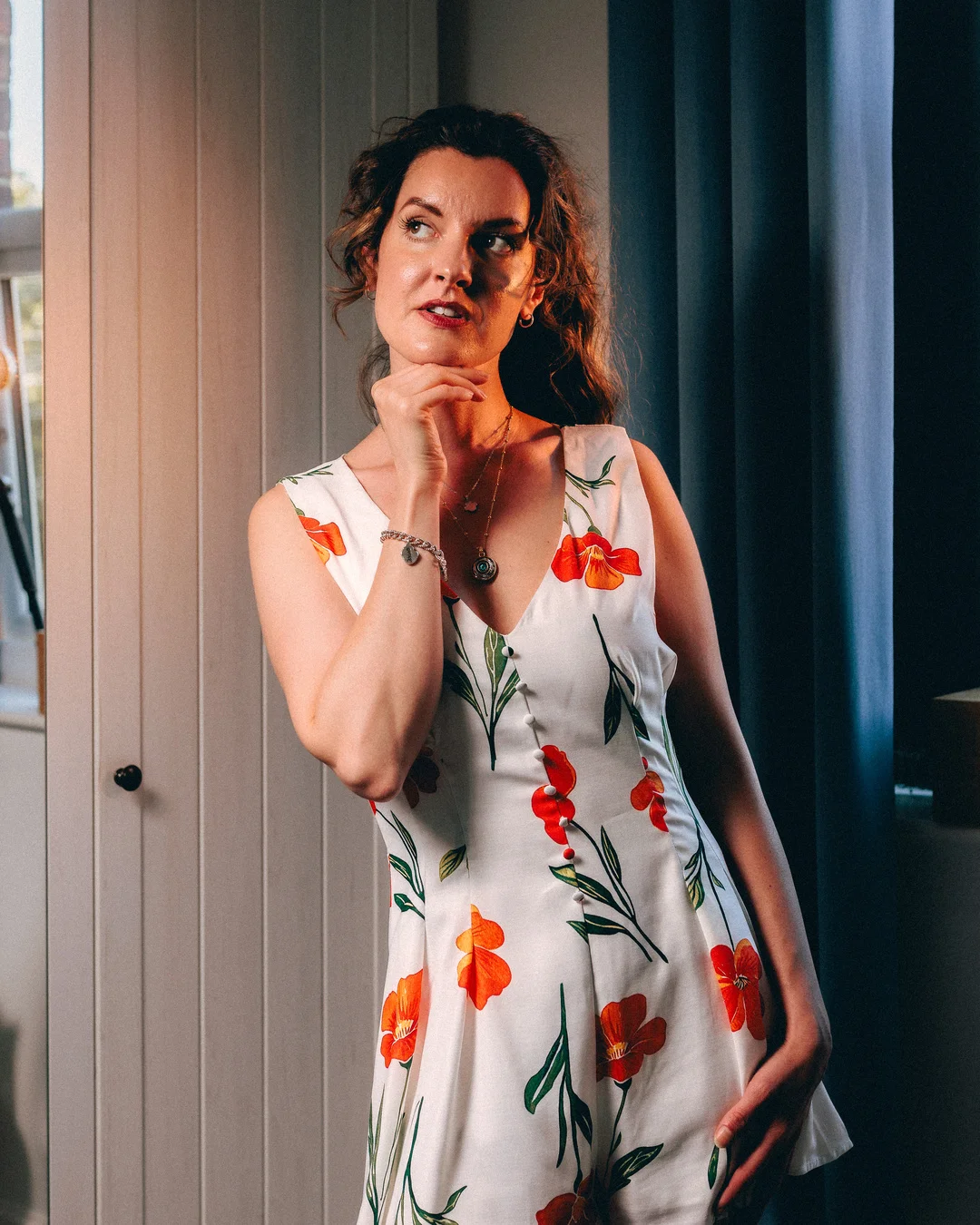 Woman in a white floral dress posing thoughtfully indoors against a white wall and blue curtain.