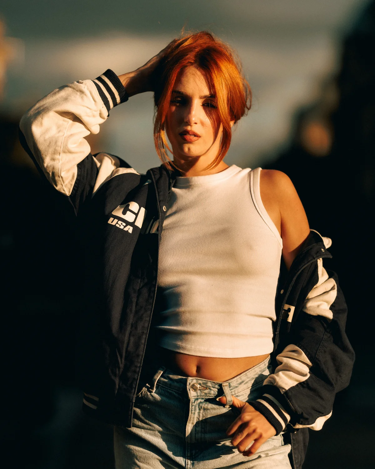 Woman with red hair posing in a varsity jacket and white crop top during golden hour.
