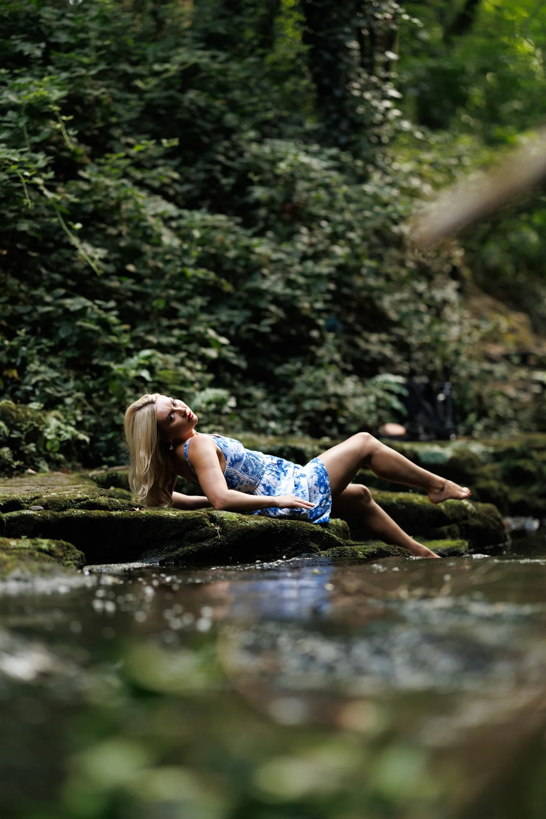 A blonde woman in a blue and white dress reclining on mossy rocks by a stream in a lush forest.
