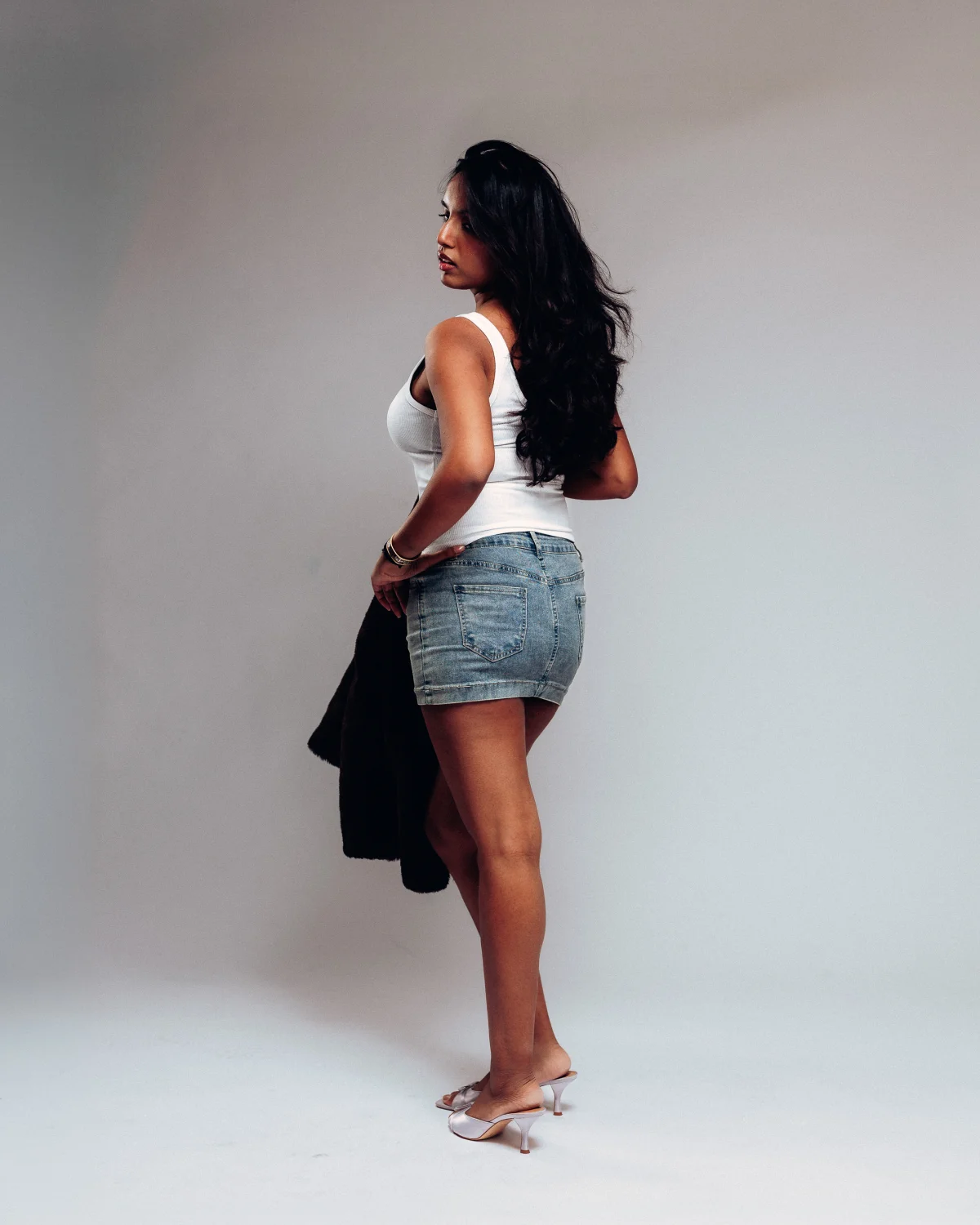 Full-body studio portrait of a woman with long dark hair wearing a white tank top and denim mini skirt.