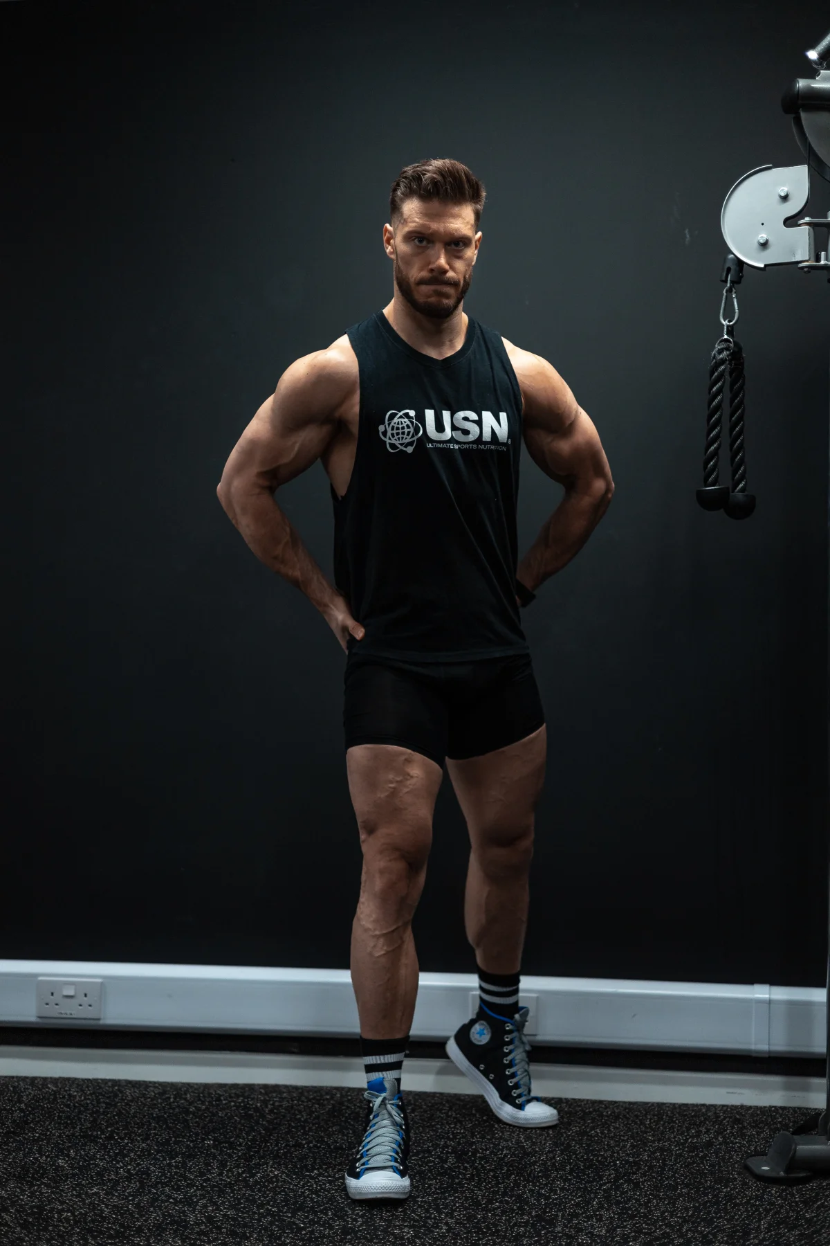Full-body portrait of a muscular man in a black tank top and athletic shorts standing in a gym against a dark wall.