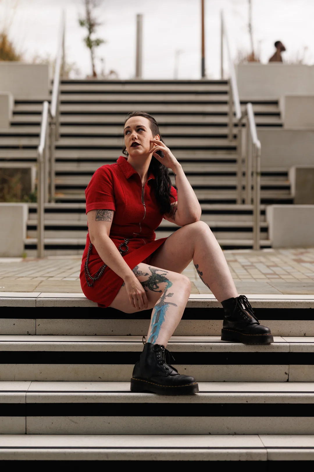 A tattooed woman in a red dress and black boots sitting on concrete outdoor stairs.
