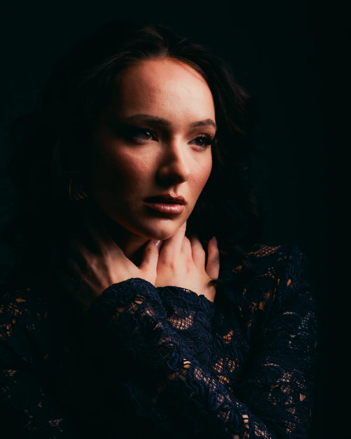 Moody close-up portrait of a woman in a dark lace dress against a black background.