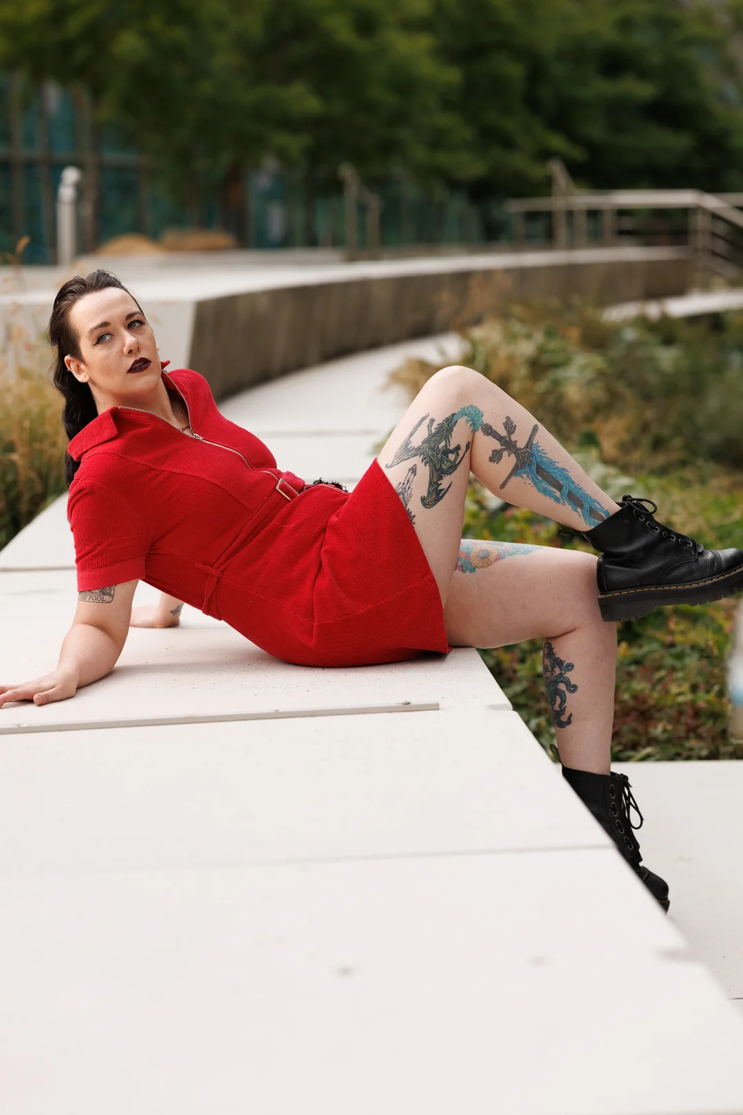 Person with tattoos in a red dress and black boots reclining on a white concrete ledge outdoors.