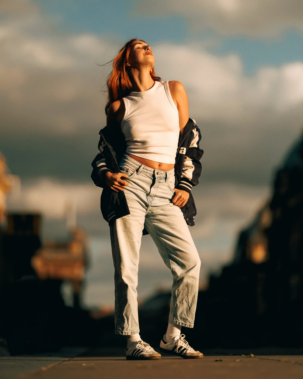 A young woman in a white tank top and light-wash jeans posing outdoors against a cloudy sky.