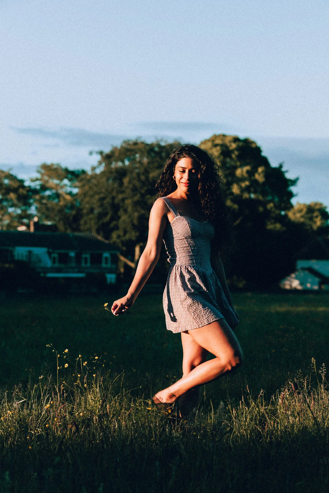 A woman with curly hair in a red and white gingham dress walking through a grassy field at sunset.