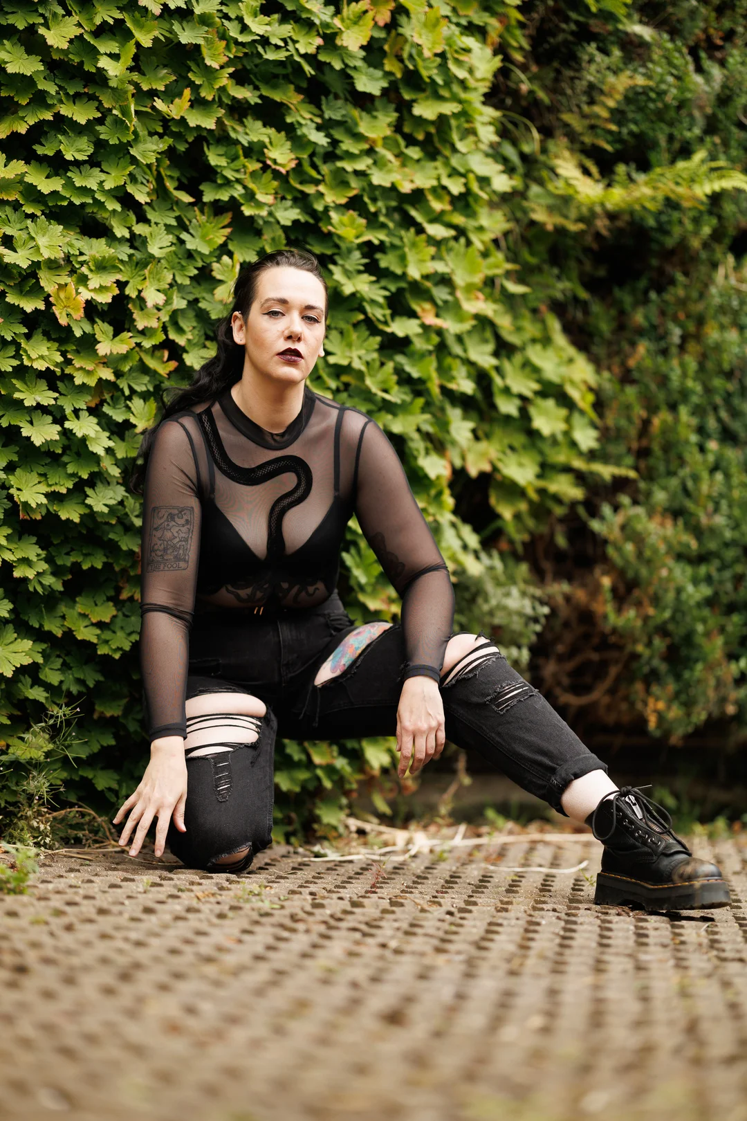Woman in a black sheer top and ripped jeans posing against a green hedge.