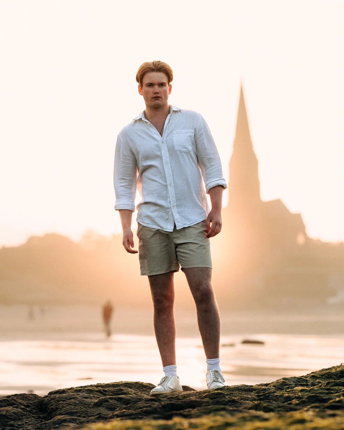 A man with red hair in a white linen shirt and khaki shorts standing on a rocky beach during golden hour.