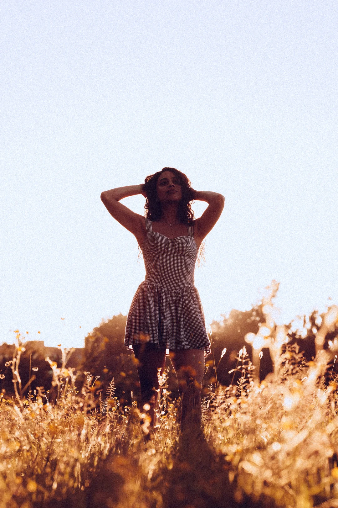 A woman in a short patterned dress standing in a golden field at sunset with her hands behind her head.