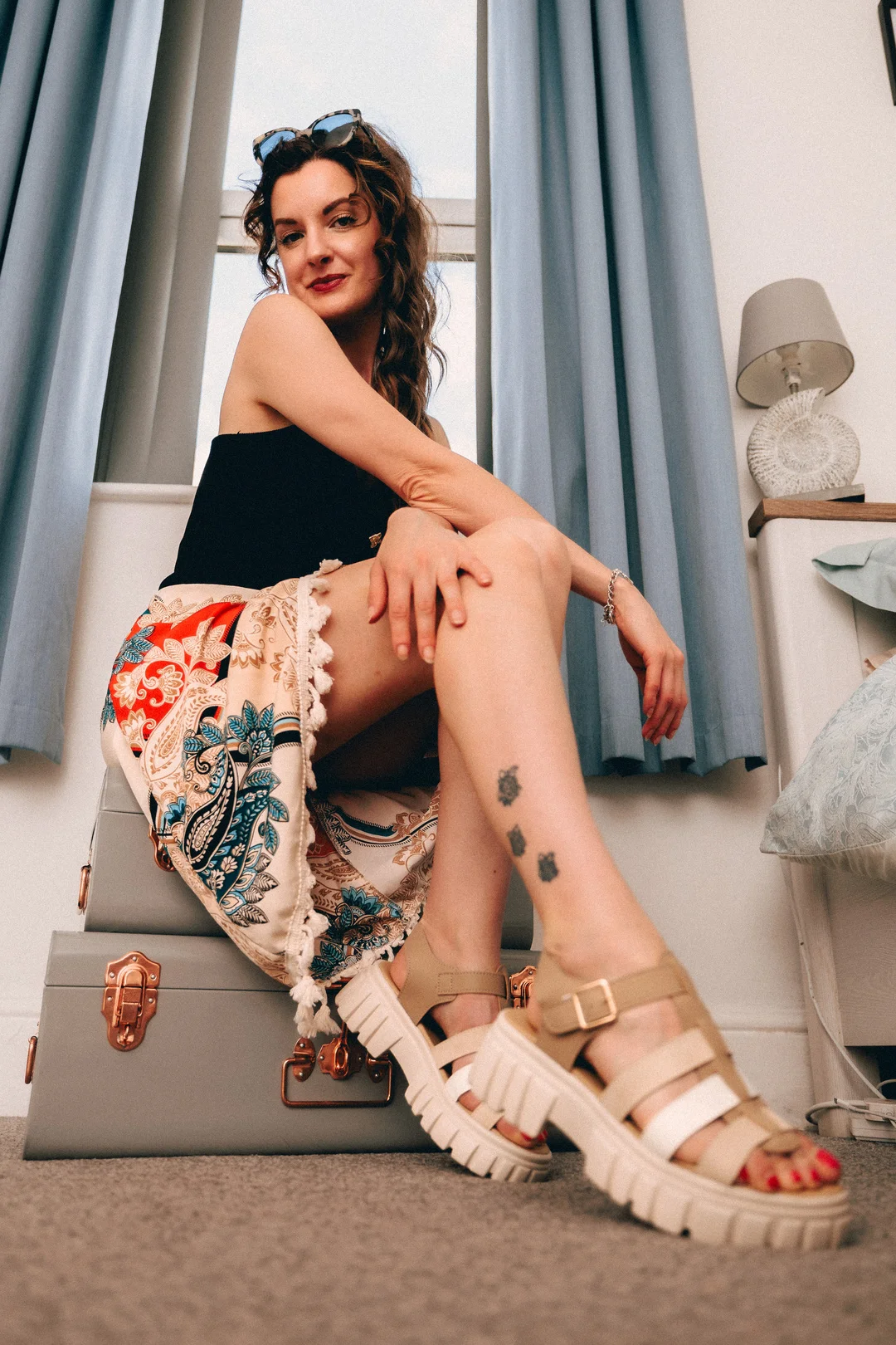 Woman in a black top and colorful patterned skirt posing on grey suitcases against blue curtains.