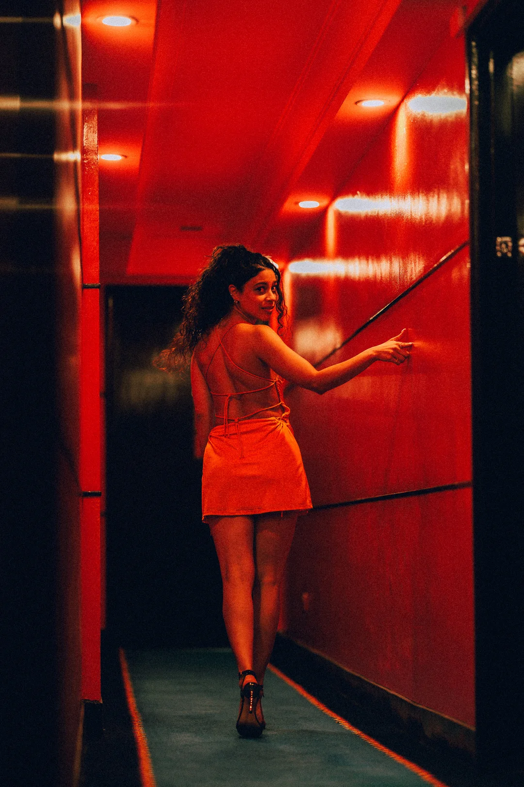 Woman in a short orange dress posing in a red-lit hallway, looking back over her shoulder.