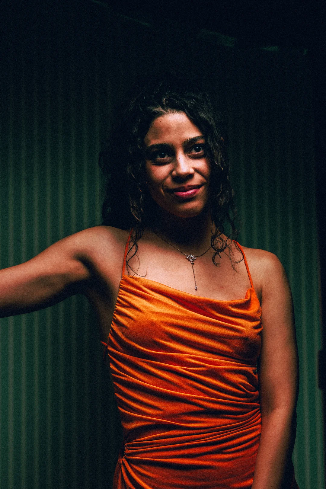 A woman with curly hair in an orange satin dress posing against a dark green striped background.