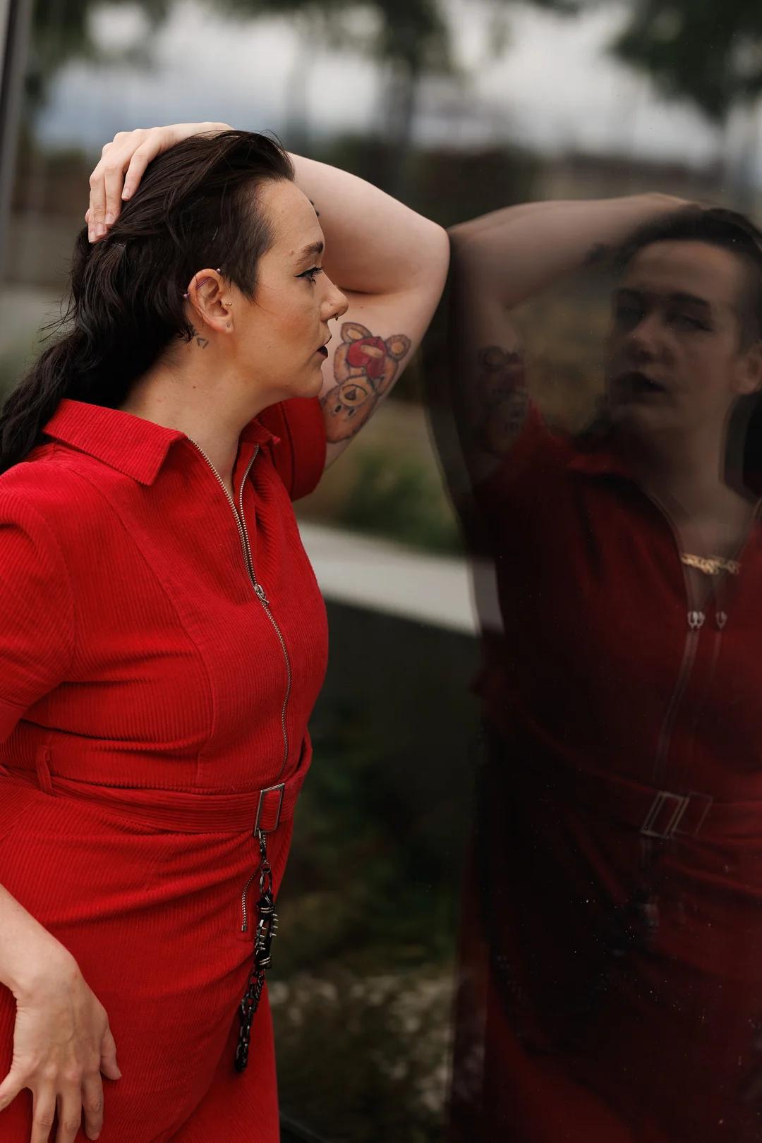 A person with dark hair and tattoos in a red corduroy outfit posing against a reflective glass surface.